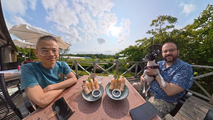 Chat, Rico y yo en el garden tailandés sentados en la terraza con dos platos de rollos frescos con gambas. El cielo es azul claro con algunas nubes y con vegetación al fondo.